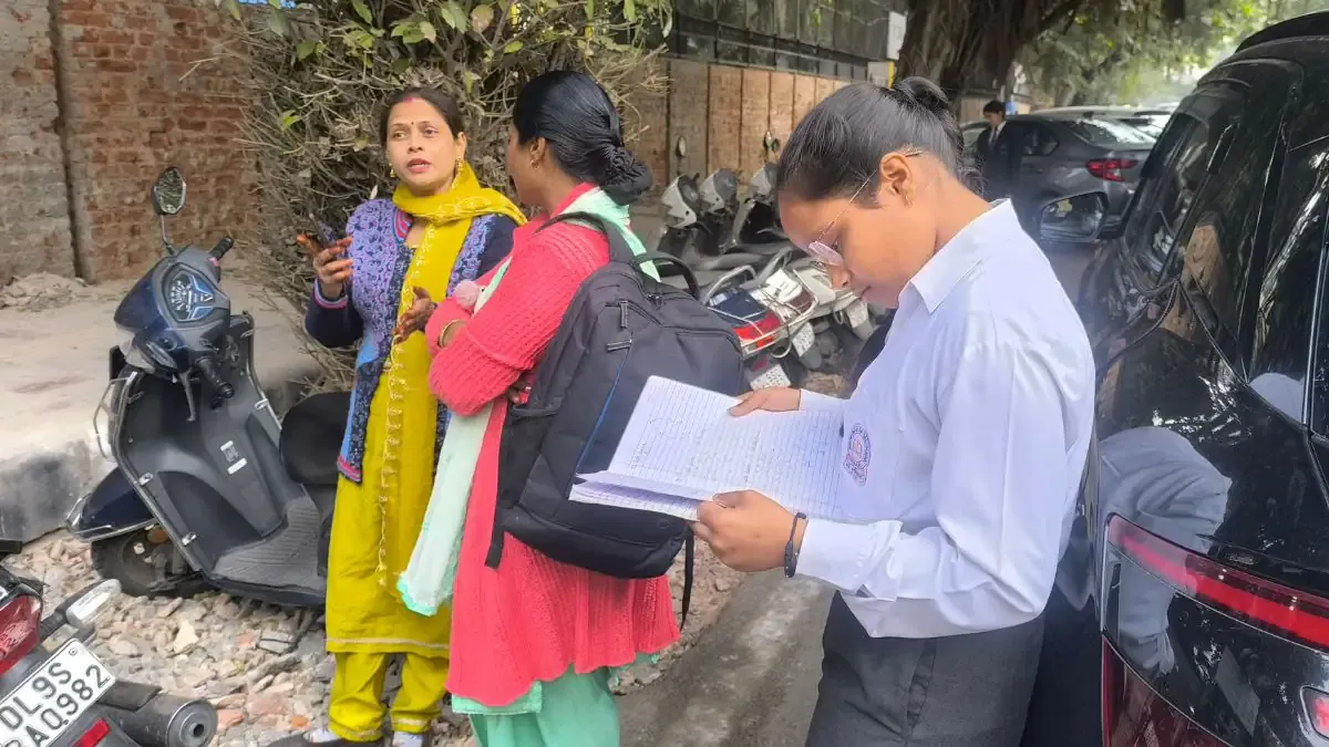 A student sitting at a desk during a CBSE board exam, focused on writing on an answer sheet with a computer in the background.
